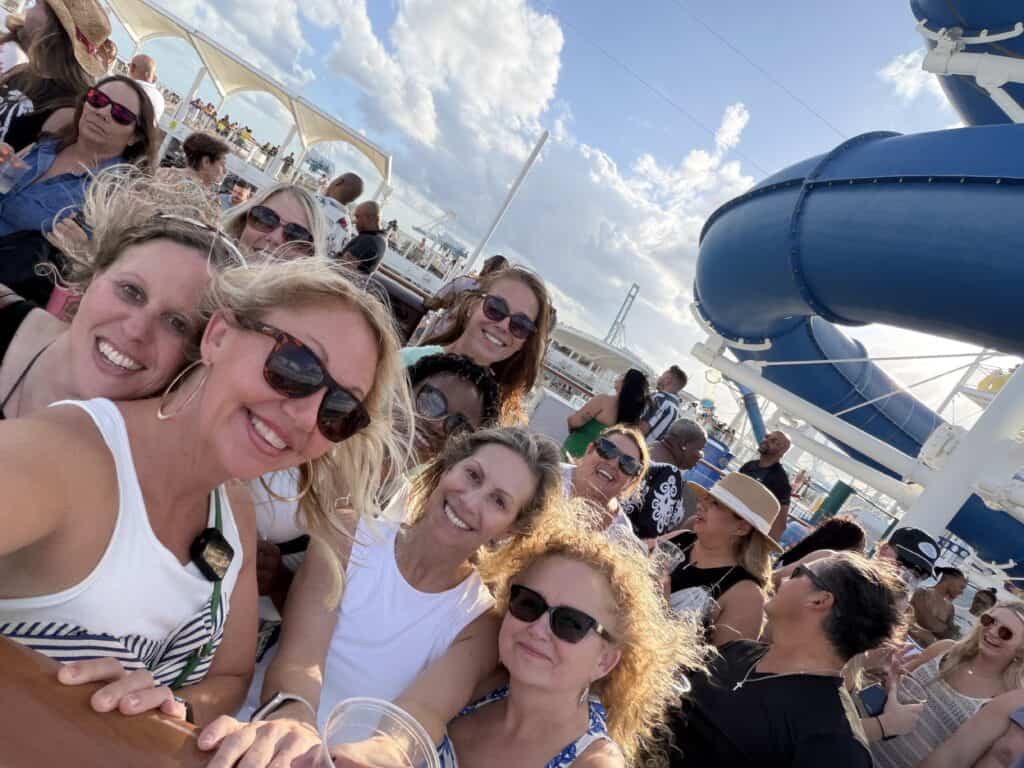 A lively group of travelers from Straight Away Travel smiles for a selfie on the sunny deck of a cruise ship. The group, mostly women wearing sunglasses and casual summer clothes, is gathered tightly together and enjoying the bright day. A large, blue water slide twists through the background on the right, under a blue sky filled with white clouds.