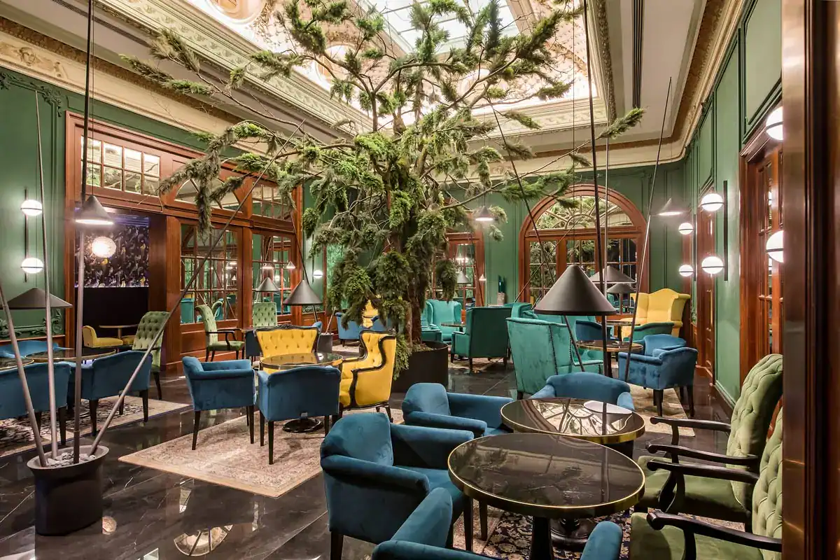 An elegant hotel lounge curated by Straight Away Travel, featuring a large decorative evergreen tree reaching toward a grand skylight ceiling in the center. The room is filled with upscale seating arrangements, including plush velvet armchairs in shades of teal, blue, yellow, and green, set around dark, polished tables. The walls are a rich forest green accented with dark wood paneling, ornate mirrors, and modern floor lamps with cone-shaped shades.