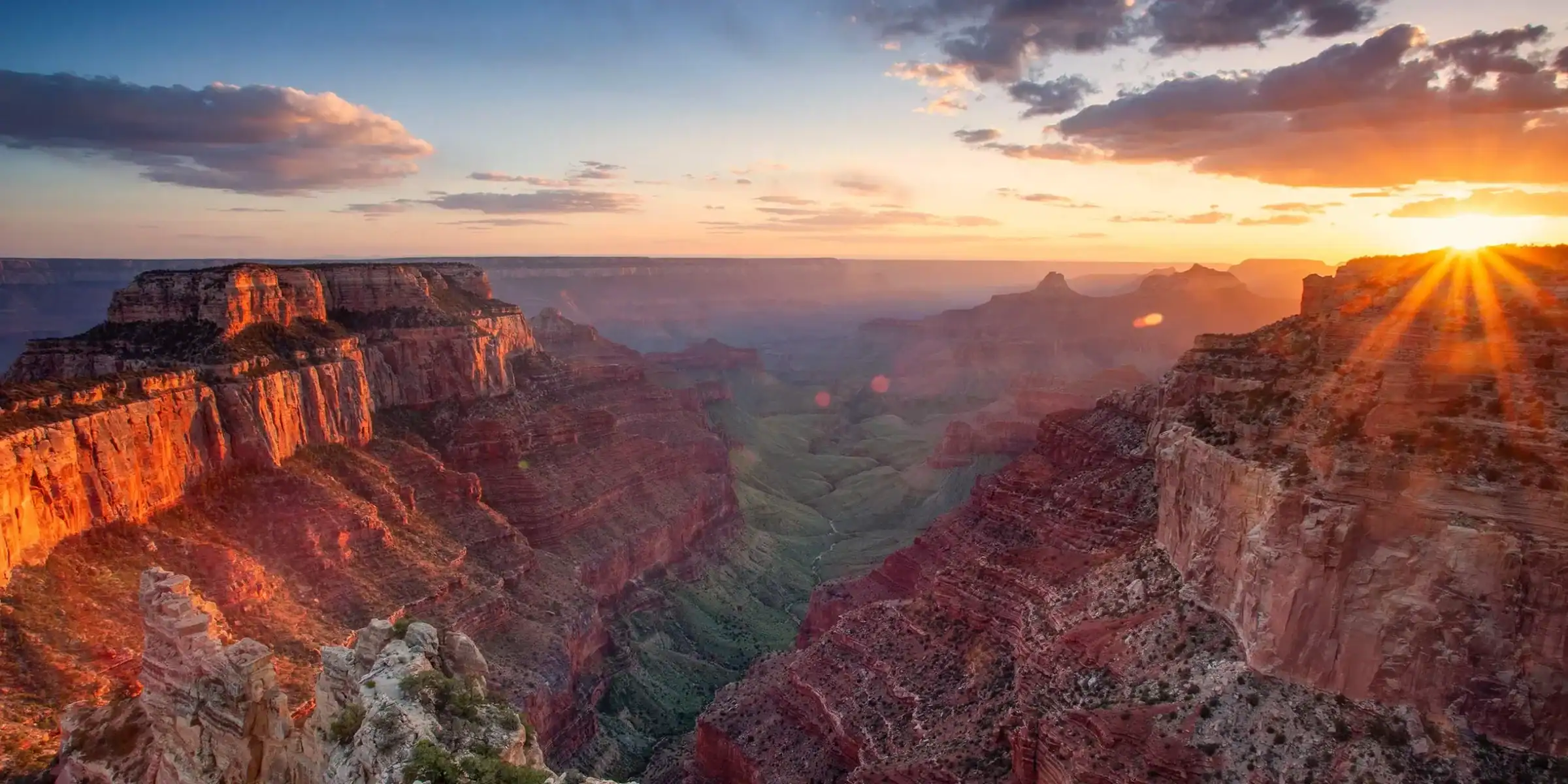 An advertisement for Straight Away Travel showcasing a panoramic view of the Grand Canyon at sunrise. The bright sun bursts from the right horizon, casting a warm golden and orange glow across the vast canyon walls, deep valleys, and layered rock formations under a softly clouded morning sky.