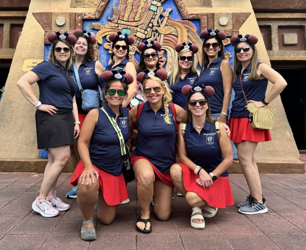 A team from Straight Away Travel poses together in matching outfits at a theme park. They are wearing dark blue sleeveless polo shirts, red skirts or black shorts, sunglasses, and Mickey Mouse ear headbands. The group consists of several women smiling for the camera, with a detailed, Aztec-style carved stone structure serving as the backdrop.