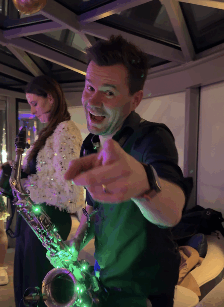 A close-up shot of a male musician performing during a live entertainment event on a Straight Away Travel vacation. He is smiling enthusiastically, looking directly at the camera, and pointing his finger forward. He holds a saxophone wrapped in glowing green fairy lights. In the background, a female performer stands under the dim, atmospheric lighting of an indoor venue with a patterned glass ceiling structure.