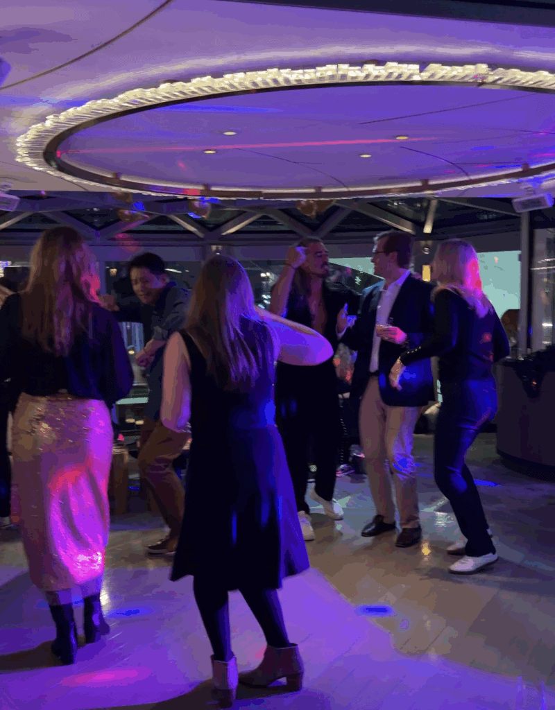 A lively group of guests on a Straight Away Travel vacation dancing together in a modern lounge or club on a cruise ship. The dance floor is illuminated by vibrant purple, pink, and blue party lights beneath a large, circular ceiling light fixture. The travelers are dressed in smart-casual and formal attire, smiling and enjoying the energetic nightlife atmosphere.