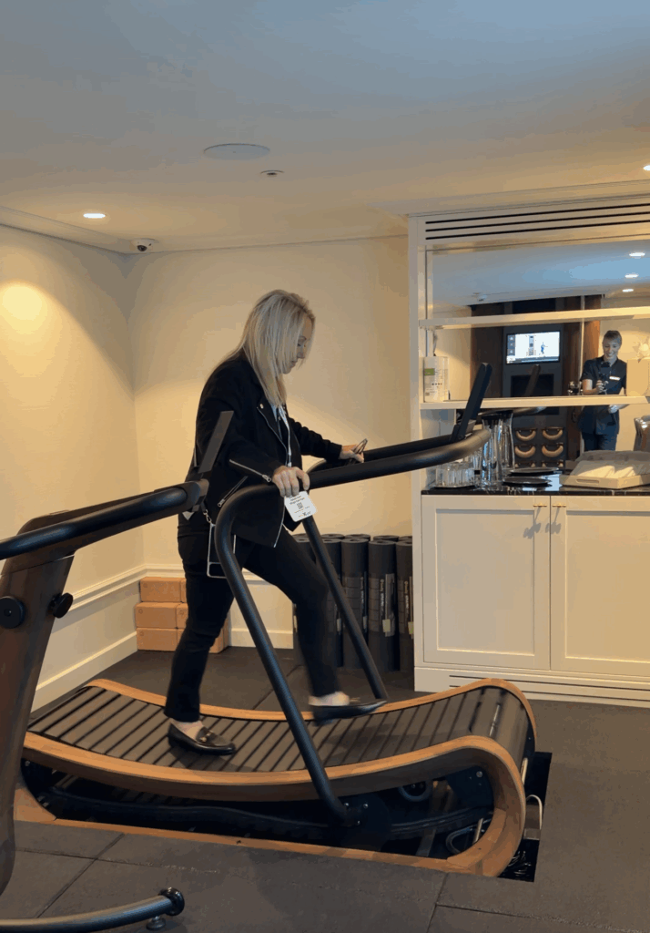 A full-length side view of a blonde woman on a Straight Away Travel vacation trying out a curved, non-motorized treadmill in a sleek indoor gym or wellness center. She is dressed in smart-casual attire, including a dark jacket, black pants, and loafers, while holding onto the treadmill's handrails. The background features a clean, white storage cabinet, rolled-up fitness mats, and a partially mirrored wall reflecting a staff member.