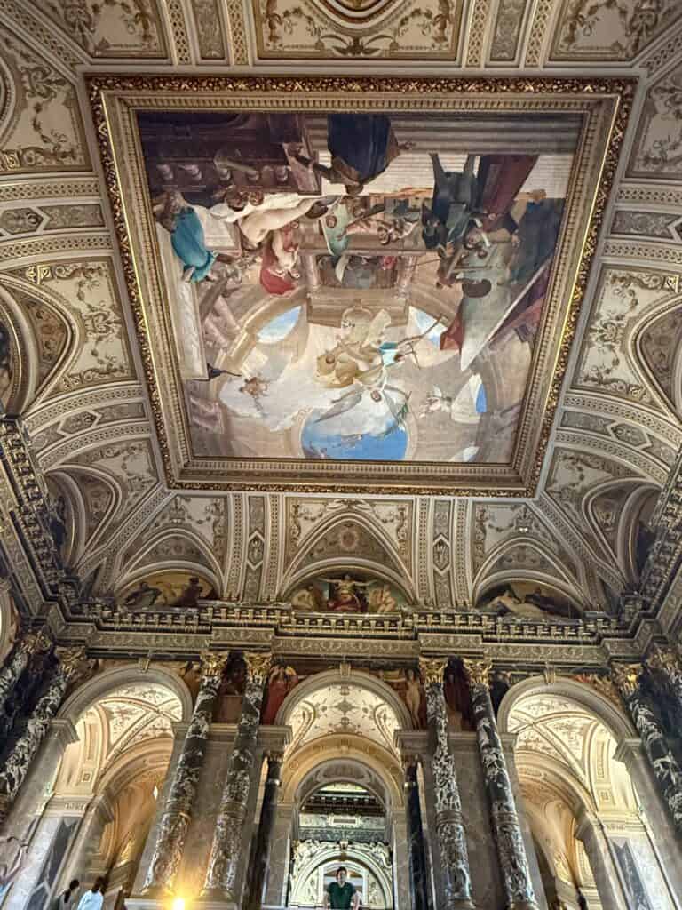 A low-angle architectural shot taken during a Straight Away Travel excursion, looking up at a grand, ornately decorated ceiling inside a historic European building. A large, classical fresco painting depicting Renaissance-style figures and a sky scene is framed in gold at the center of the vaulted ceiling. The surrounding structure features intricate gold molding, detailed archways, and massive, dark marble columns that frame the opulent hall below.