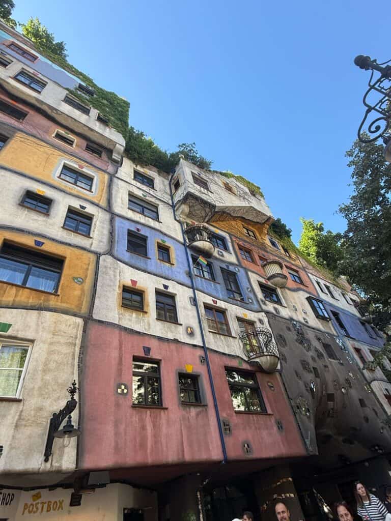 A low-angle shot of the famous Hundertwasserhaus in Vienna, capturing its whimsical, non-linear architecture under a clear blue sky. The facade features a patchwork of vibrant colors—including pink, blue, yellow, and white—with uneven windows, integrated greenery growing from the balconies, and wavy lines instead of straight edges. At the very bottom left, a sign for a postbox is partially visible, while the foliage of trees framing the right side hints at an immediate straight away travel destination.