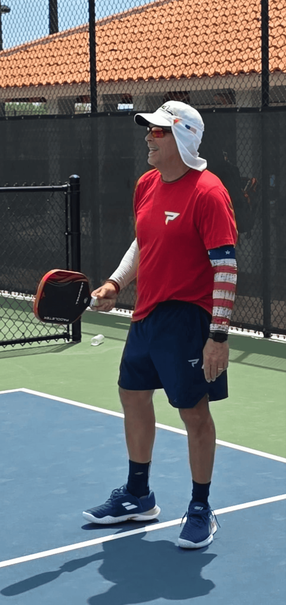A representative from Straight Away Travel stands on an outdoor pickleball court during a sunny day, holding a paddle and a ball. He is wearing a red athletic t-shirt, navy blue shorts, blue tennis shoes, sunglasses, and a white sun hat with a neck flap. He also wears patriotic, American flag-themed compression sleeves on his arms while looking off to the side of the blue and green court.