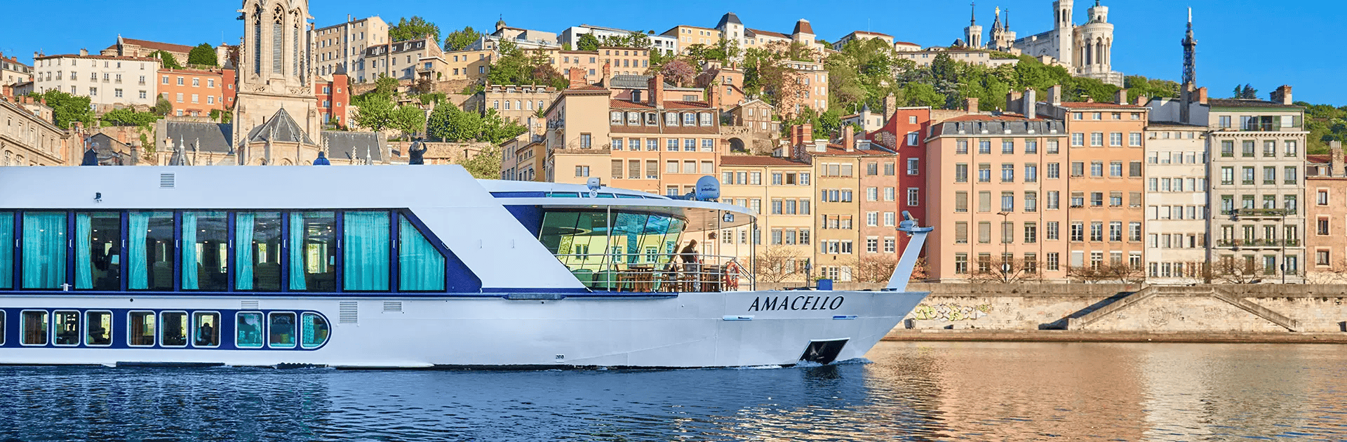 A scenic travel destination documented by Straight Away Travel features a white and blue river cruise ship named "AMACELLO" gliding along a wide river. In the background, a historic European city rises up a hillside, showcasing tightly packed, multi-story buildings in warm pastel tones, old church steeples, and lush green trees under a clear blue sky.