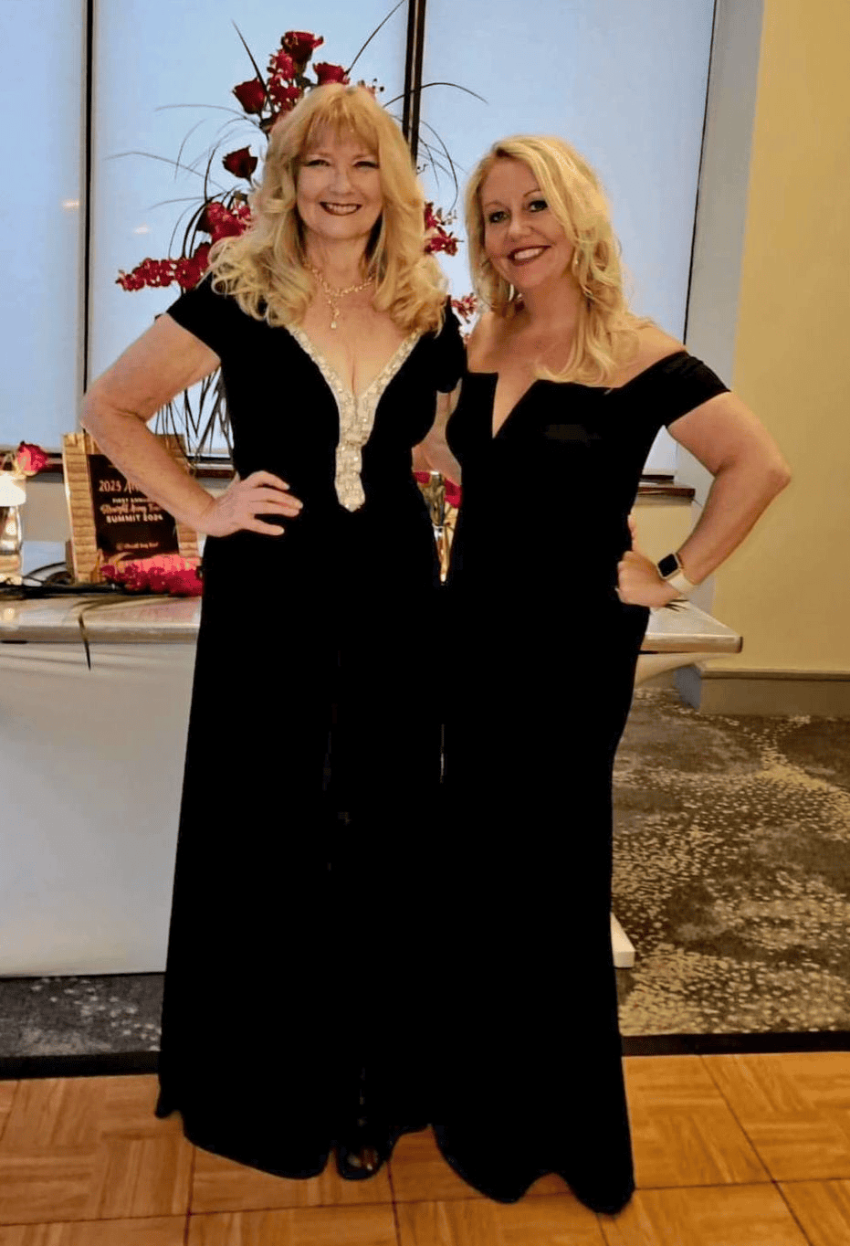 Two elegant professionals representing Straight Away Travel smile for a photo at a formal event. Both blonde women are wearing long, sophisticated black evening gowns and standing on a wooden dance floor. The woman on the left wears a gown with a beaded, plunge neckline, while the woman on the right wears an off-the-shoulder style dress. A decorative floral arrangement and an award plaque are visible on a table in the background.