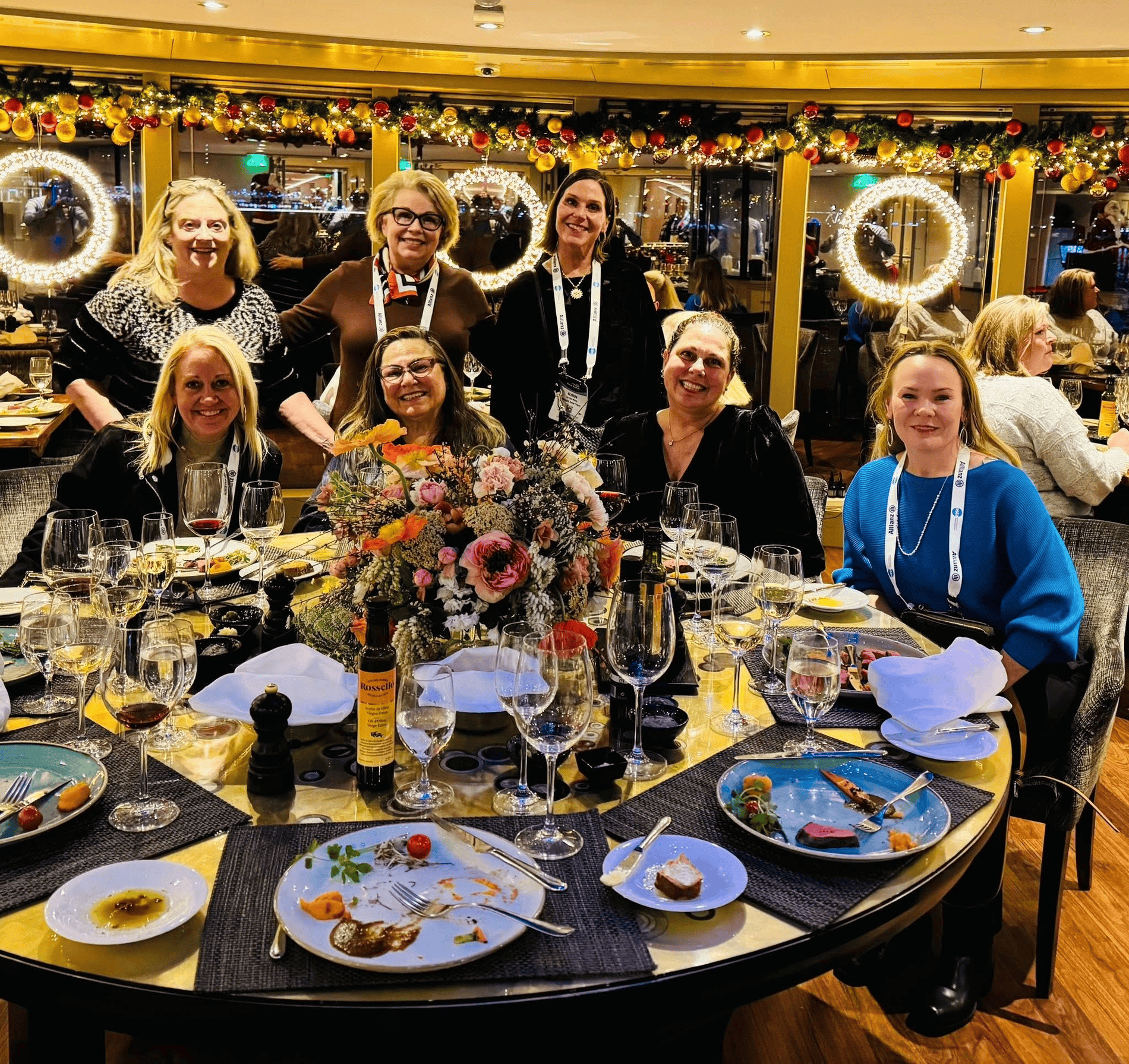 A group of travel professionals representing Straight Away Travel smiles together around a large, round dinner table at a festive restaurant. The group of seven women is gathered closely, some seated and some standing, during a formal or holiday dinner. The table is elegantly set with a large floral centerpiece, multiple wine glasses, and plates with the remains of a meal. The background features warm, golden lighting with festive holiday garlands and glowing circular wreaths.