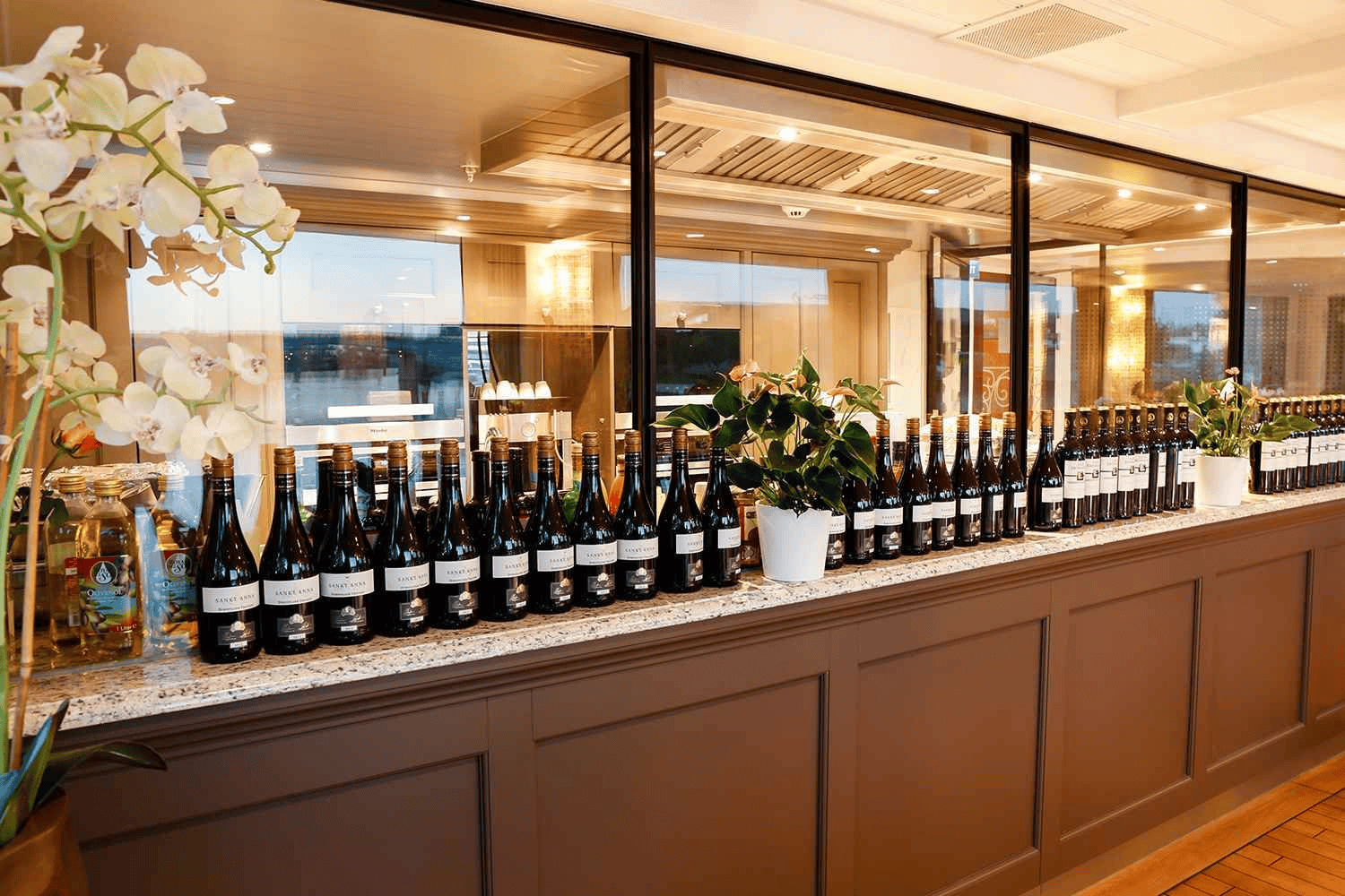 A luxury cruise ship dining experience, documented by Straight Away Travel, features a long counter elegantly lined with dozens of identical wine bottles with dark glass and white labels. The granite-topped counter sits beneath a large glass partition showing a glimpse of the kitchen or dining area behind it. The sophisticated setup is accented by potted green plants and a white orchid floral arrangement on the left under warm, bright lighting.