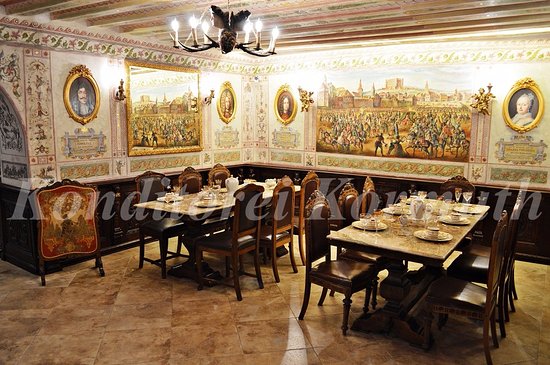 An interior view of a richly decorated dining room featuring two large stone-topped tables set with porcelain teacups and matching high-backed leather chairs. The walls are adorned with ornate murals depicting historical panoramic scenes, framed portrait paintings, and decorative borders. A rustic antler-style chandelier hangs from the exposed-beam ceiling, illuminating the warm, tiled flooring.
