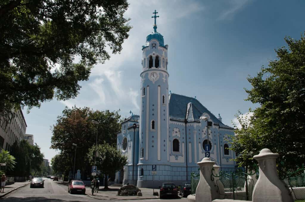 The exterior of a striking Art Nouveau church painted in bright light blue with white trim, featuring a tall, cylindrical bell tower with an integrated clock and a blue dome topped with a cross. The building has curved architectural details and a blue-tiled roof, set against a partly cloudy sky with trees and parked cars visible at the street level.