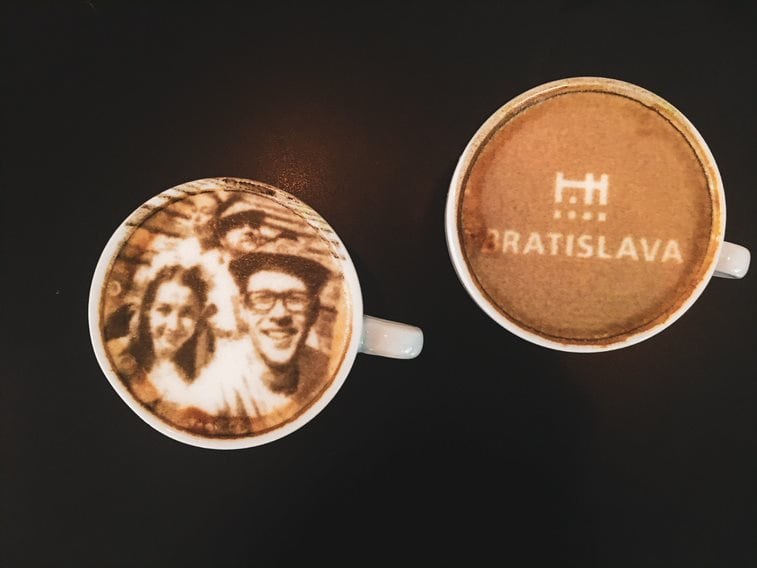 A top-down view of two coffee cups on a dark background, featuring custom latte art. The cup on the left displays a highly detailed portrait of a smiling couple printed directly onto the foam. The cup on the right features a white printed design showing a minimalist castle icon and the text "BRATISLAVA" neatly stenciled across the surface.